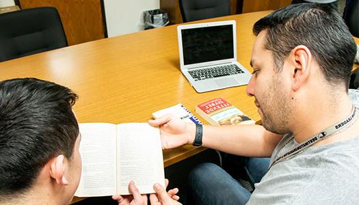 a student and instructor sit at a table with a laptop looking at a book